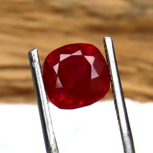 May include: A vibrant, cushion-cut ruby gemstone held in silver tweezers. The ruby displays a rich, deep red hue, with internal light reflections. The background is a blurred, neutral tone, emphasizing the gemstone's clarity and colour.