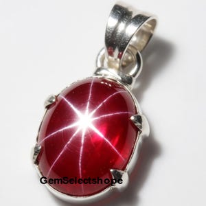 Puede incluir: Colgante de piedra preciosa roja pulida, de forma ovalada, con un patrón en forma de estrella. La piedra preciosa está engastada en un metal plateado con un lazo para una cadena. El texto "GemSelectshope" es visible en la parte inferior del colgante.