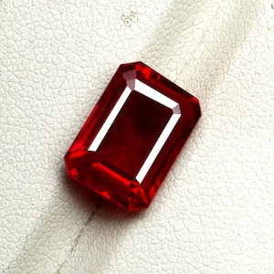 Pode incluir: Uma gema vermelha escura facetada, com corte retangular, com um interior branco brilhante. A gema está sobre uma superfície texturizada esbranquiçada, mostrando sua clareza e cor. A forma da gema é um corte esmeralda.