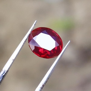 Pode incluir: Uma gema de rubi facetada, em forma oval, segurada por pinças prateadas. O rubi é de uma cor vermelha profunda e rica, com uma superfície brilhante e reflexiva. A gema é cortada para maximizar a reflexão da luz.