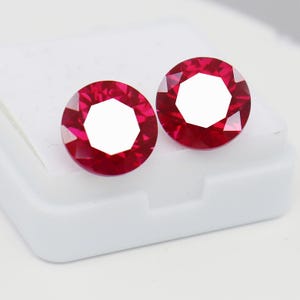 Könnte beinhalten: Zwei runde, facettierte, leuchtend rote Edelsteine im Brillantschliff mit klaren geometrischen Facetten und einer hellen, reflektierenden Oberfläche. Diese losen Steine liegen nebeneinander auf einem schlichten, quadratischen weißen Sockel.