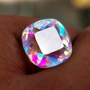 May include: A large, square-cut gemstone with a rainbow iridescence. The gem has a clear, central square and facets that reflect a spectrum of colors, including pink, blue, and yellow. The gem is held in a hand.