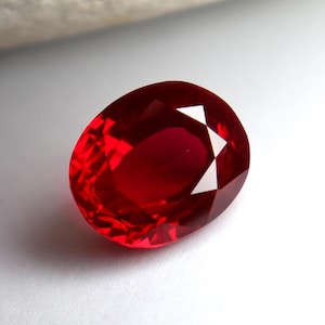 Peut inclure: Une pierre précieuse de forme ovale, un rubis. La gemme est d'un rouge profond et saturé, avec de multiples facettes qui reflètent la lumière. Le rubis est posé sur un fond blanc, ce qui met en valeur son éclat et sa clarté.