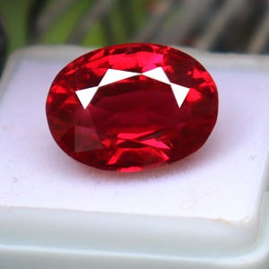 May include: A vibrant, oval-cut gemstone with a deep, saturated red color and multiple facets that reflect light. The stone is displayed on a white surface, showcasing its transparent clarity and polished finish with distinct geometric light reflections.