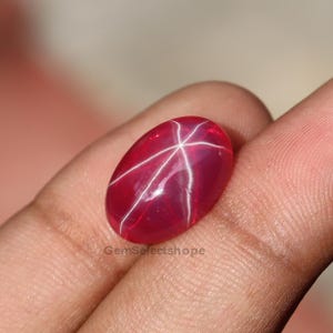 May include: A polished, oval-shaped, red gemstone with a six-rayed star pattern. The star effect is created by white, intersecting lines across the surface of the gem. The gemstone is held in a hand, with the text "GemSelectshope" visible.