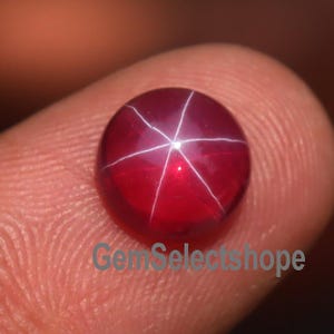 Könnte beinhalten: Ein runder, polierter, tiefroter Edelstein mit einem sechsstrahligen Sternmuster. Der Edelstein hat eine glatte, reflektierende Oberfläche. Der Text "GemSelectshop" ist unten im Bild sichtbar.