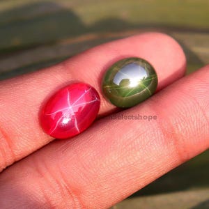 Puede incluir: Dos gemas ovaladas. Una es de color rojo vibrante con un patrón de estrella de seis rayos, la otra es verde oscuro con un efecto de estrella similar. Las gemas se sostienen en una mano, mostrando sus superficies pulidas.