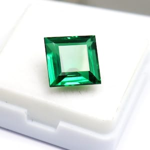 May include: A square-cut, faceted gemstone with a vibrant, translucent emerald green color. The stone features a clear, polished surface with sharp edges and a symmetrical geometric shape, displaying internal light reflections.