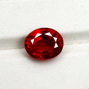 May include: A vibrant, oval-cut ruby gemstone. The deep red stone has facets that reflect light, creating a sparkling effect. The gem is set against a white, textured background, highlighting its rich color and brilliance.
