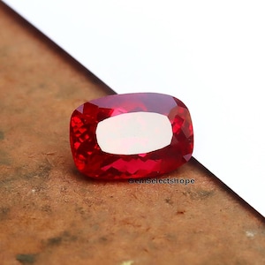 May include: A faceted, cushion-cut, red gemstone with a bright, reflective surface. The gem has a large, oval-shaped, white inclusion in the centre. The text "GemSelectshope" is visible on the surface.