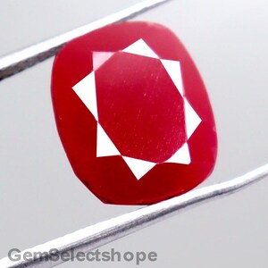 May include: A vibrant red gemstone with a square cut and a star-like pattern in the center. The gem is held by silver tweezers against a white background. The text "GemSelectshope" is visible at the bottom.