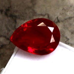 Può includere: Una gemma sfaccettata a forma di goccia, di colore rosso intenso. La gemma ha una superficie lucida con molteplici sfaccettature che riflettono la luce, creando riflessi brillanti. La gemma è posta su una superficie bianca.