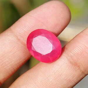 May include: A faceted, oval-shaped ruby gemstone. The gem is a vibrant shade of red with a polished surface, showcasing internal facets that reflect light. The ruby is held between fingers, with a blurred background.