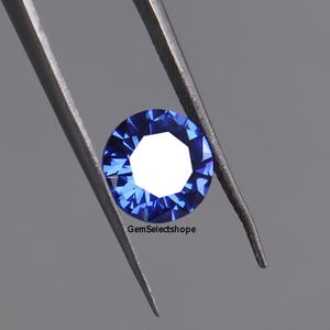 Peut inclure: Une pierre précieuse saphir ronde et facettée tenue par une pince. Le saphir est d'un bleu profond avec un centre blanc. La pierre précieuse est ronde et comporte de nombreuses facettes, reflétant la lumière. Le texte "GemSelectshope" est visible.