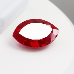 Puede incluir: Una piedra preciosa de talla marquesa con un color rojo intenso y vibrante, con múltiples superficies facetadas que reflejan la luz. La piedra tiene una forma ovalada puntiaguda con lados simétricos y un aspecto pulido y translúcido.