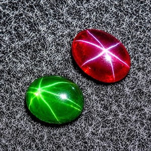 以下が含まれることがあります： 鮮やかな赤と深い緑の2つの楕円形のカボションカットの宝石。それぞれにアステリズムと呼ばれる、光を反射して浮かび上がる6条の星のような模様が見られます。石の表面は滑らかで光沢があり、光を反射して白い星の形を作り出しています。
