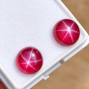 Könnte beinhalten: Zwei runde, im Cabochon-Schliff gearbeitete Sternrubine in tiefroter Farbe mit einem deutlichen, sechsstrahligen Asterismus-Effekt auf der Oberfläche jedes Edelsteins. Die Steine liegen nebeneinander in einer weißen, rechteckigen Schmuckschatulle.