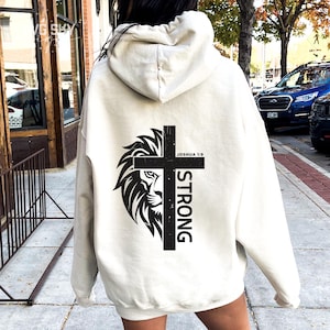 Puede incluir: Sudadera con capucha color crema con un gráfico negro en la espalda. El gráfico presenta la cabeza de un león, una cruz y la palabra "STRONG" en orientación vertical. También incluye el texto "JOSHUA 1:9".
