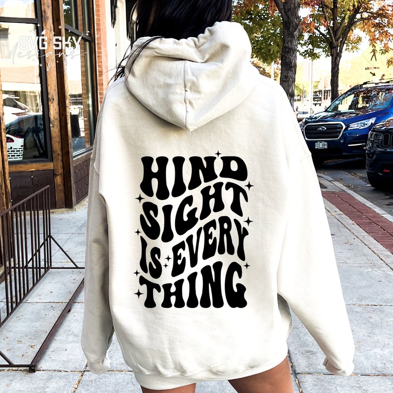May include: A white hoodie with a black graphic print on the back that says "Hind Sight Is Every Thing" in a retro font.