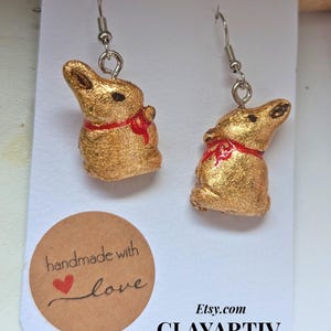Könnte beinhalten: Ein Paar goldfarbene Hasenohrringe mit roten Schleifen. Die Ohrringe sind handgefertigt und haben eine strukturierte Oberfläche. Das Bild zeigt auch einen Aufkleber mit der Aufschrift "handmade with love" und den Etsy-Shop-Namen.