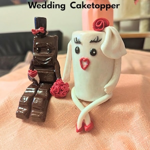 Könnte beinhalten: Ein skurriler Tortenaufsatz für Hochzeiten mit einem Schokoriegel-Bräutigam mit Zylinder und einer weißen Milchkarton-Braut. Braut und Bräutigam sind mit roten Rosen und Schleifen geschmückt. Der Text "Schocki & Milky Wedding Caketopper" ist zu sehen.