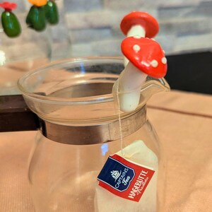 Könnte beinhalten: Eine transparente Glasteekanne mit einem pilzförmigen Tee-Infuser. Der Pilz hat einen roten Hut mit weißen Punkten. Ein Teebeutel mit einem blau-roten Etikett befindet sich in der Teekanne. Der Etsy-Shop-Name "CLAYARTIV" steht unten.
