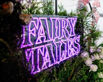 May include: A neon sign with the words "FAIRY TALES" in bright purple. The sign is surrounded by greenery, white flowers, and decorative playing cards. The sign is a decorative element.