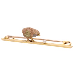Op de afbeelding: Een gouden broche met een klein vogeltje dat op een staafje zit en naar een parel kijkt. Het vogeltje is gedetailleerd met emaille en heeft rode ogen.