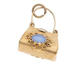 May include: A gold charm in the shape of a handbag with a blue gemstone in the center. The handbag has a detailed floral design around the gemstone.