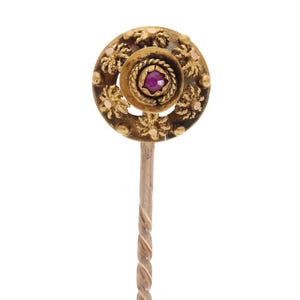 May include: A gold-toned stick pin with a circular head. The head features a central, round, red gemstone surrounded by a detailed, floral-like design. The pin's stem has a twisted rope design.