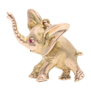 May include: A gold-colored elephant pendant with a raised trunk and a red gemstone eye. The pendant is detailed with textured surfaces and a loop for attaching to a chain or bracelet. The elephant is standing on four legs.