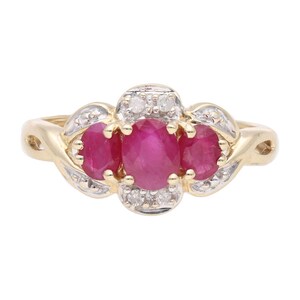Op de afbeelding: Een gouden ring met drie ovale, helderrode edelstenen, waarschijnlijk robijnen, in een clusterontwerp. De ring is versierd met kleinere, heldere edelstenen, mogelijk diamanten, en heeft een sierlijke band.