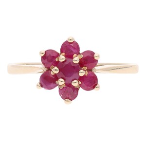 May include: A gold ring featuring a flower design with seven round, deep red gemstones. The gemstones are set closely together, forming a floral pattern. The ring's band is a simple, polished gold.