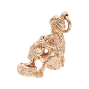 May include: A gold-colored charm depicting a seated figure playing a drum. The charm has a loop at the top for attaching to a bracelet or necklace. The figure is detailed with facial features and clothing.
