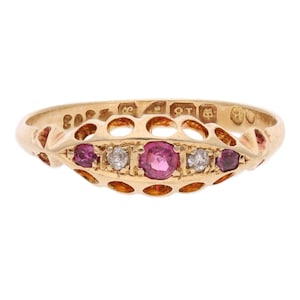 May include: A gold ring featuring a central row of three round pink gemstones and two round clear gemstones set within a decorative openwork gallery. The interior of the band displays engraved hallmarks including the number 18.