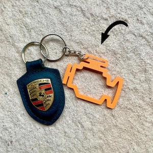 May include: A keychain set with a navy blue leather tag bearing the Porsche emblem and an orange engine-shaped charm. The Porsche emblem is gold with red and black details. The keychain has a silver metal ring.