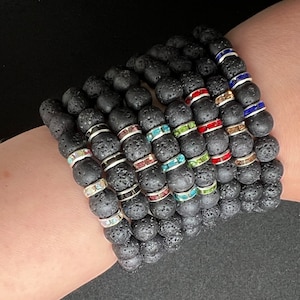 May include: Several black lava stone bead bracelets with colourful rhinestone accents. The bracelets have silver, blue, red, green, and clear rhinestones. The bracelets are worn on a wrist.