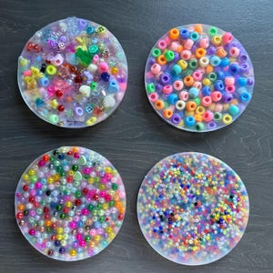 May include: Four circular resin coasters, each embedded with colourful beads and small embellishments. One coaster has a mix of beads and charms, another has orange, purple, and green beads, a third has pearls, and the last has a mix of small beads.