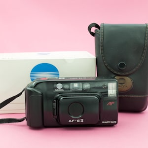 May include: A black Minolta AF-E II camera with a black leather case. The camera has a silver lens and a silver button on the top. The camera is on a pink surface.