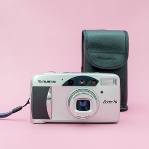 May include: A silver Fujifilm Zoom 70 point-and-shoot camera with a black leather case. The camera has a lens with a zoom range of 38-70mm and an auto focus feature.