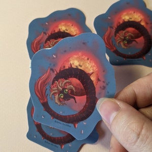 Puede incluir: Una pegatina con una criatura roja y negra con grandes ojos y espinas, enroscada alrededor de un sol rojo y naranja sobre un fondo azul con motas blancas. La pegatina tiene el texto "Selene.the.blue" en la parte inferior.