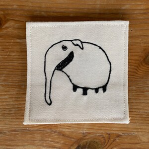 Könnte beinhalten: Ein weißer Stoffuntersetzer mit einem schwarzen gestickten Elefantenmotiv. Der Elefant hat einen langen Rüssel und ein kleines Auge.
