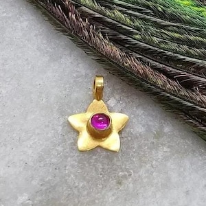 May include: A gold star-shaped pendant with a pink gemstone in the centre. The pendant is on a white marble surface.