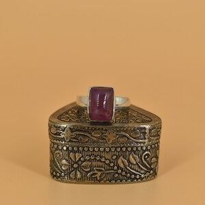 May include: A silver ring with a rectangular, purple gemstone set in the center. The ring is sitting on top of a silver, ornate box.