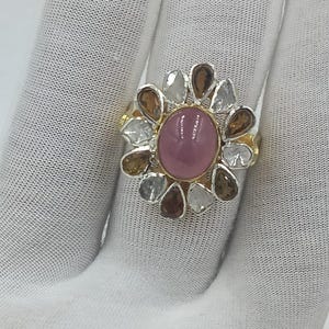 May include: A gold and silver ring featuring a large oval pink gemstone at the center, surrounded by a floral arrangement of smaller, multi-colored gemstones. The ring is set on a white surface.