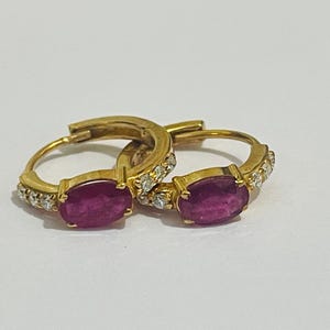 Puede incluir: Un par de pendientes de aro dorados. Cada pendiente presenta una gran gema de rubí rojo oscuro de forma ovalada. Los aros también están adornados con pequeñas gemas transparentes. Los pendientes se muestran sobre una superficie blanca.