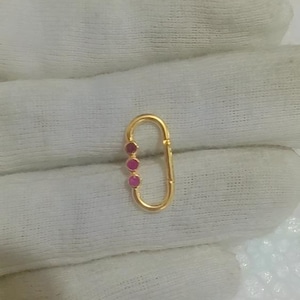 May include: A gold-colored, oval-shaped clasp with three small, round, red gemstones set along one side. The clasp is shown against a white, textured background, suggesting a jewelry or accessory item.