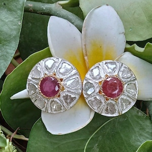 May include: A pair of silver earrings with a round design. Each earring features a red ruby surrounded by small diamonds.