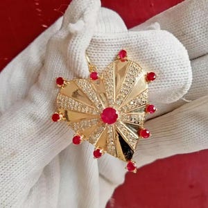 Puede incluir: Un colgante de corazón de oro con un diseño de sol y piedras preciosas rojas alrededor del borde.