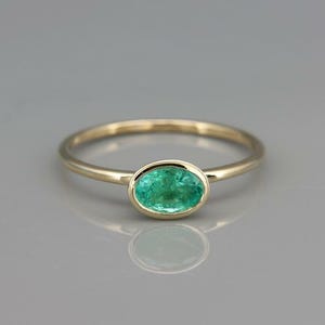 Puede incluir: Un anillo de oro con una gema verde ovalada vibrante. El anillo tiene una banda delgada y pulida, y la gema está engastada en un bisel. El anillo se muestra sobre una superficie reflectante.